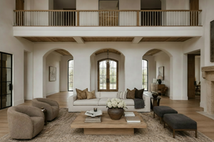 A high-ceilinged, open-plan living room with arched entryways, wood floors, a white sofa with brown and beige pillows, a wooden coffee table with flowers, two upholstered armchairs, and dark gray ottomans. A second-floor balcony with a wood railing is visible above.